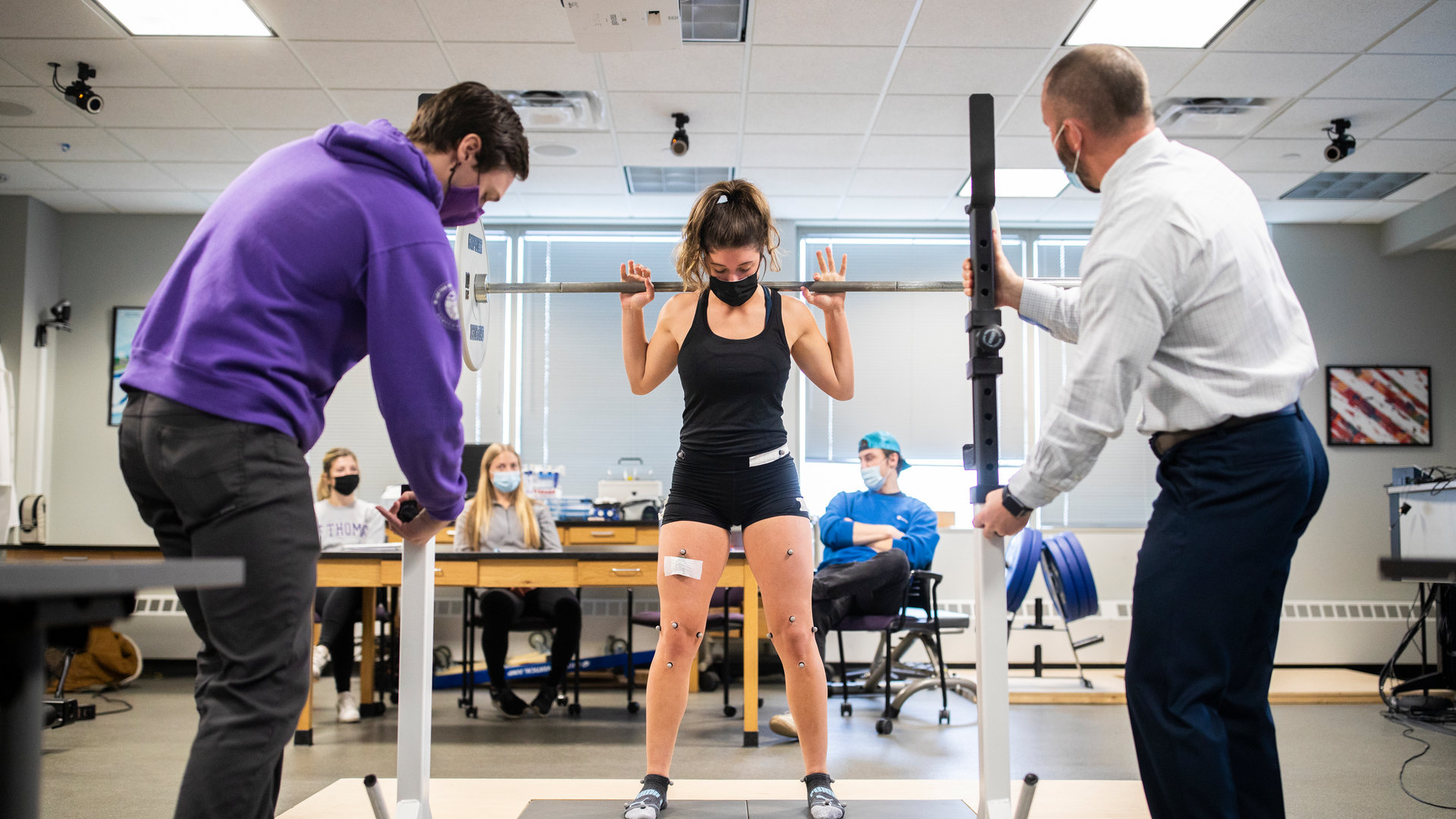 Professor and student observe another student wearing biometric sensors while doing squats.
