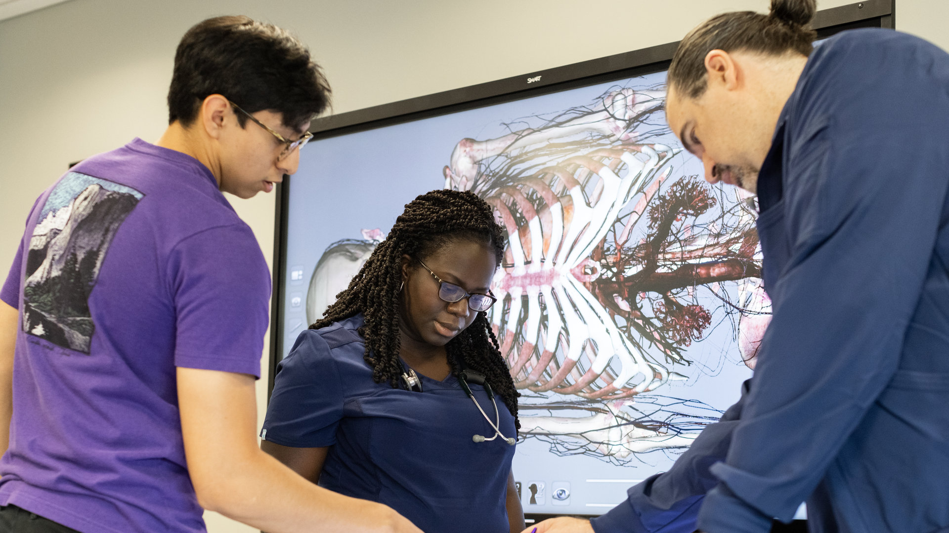 A student and two faculty interact with a digital anatomy tool in a lab classroom. 