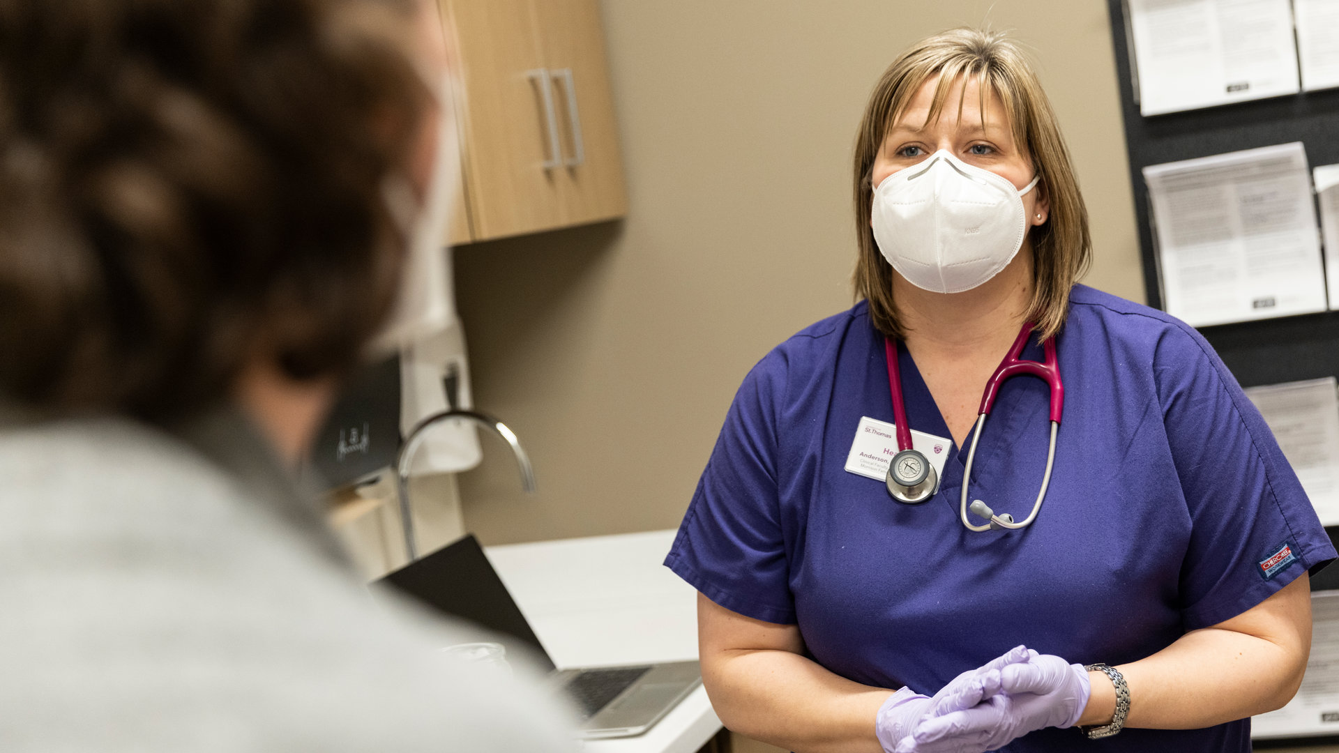A female nurse in a mask speaks with a patient in a medical office.