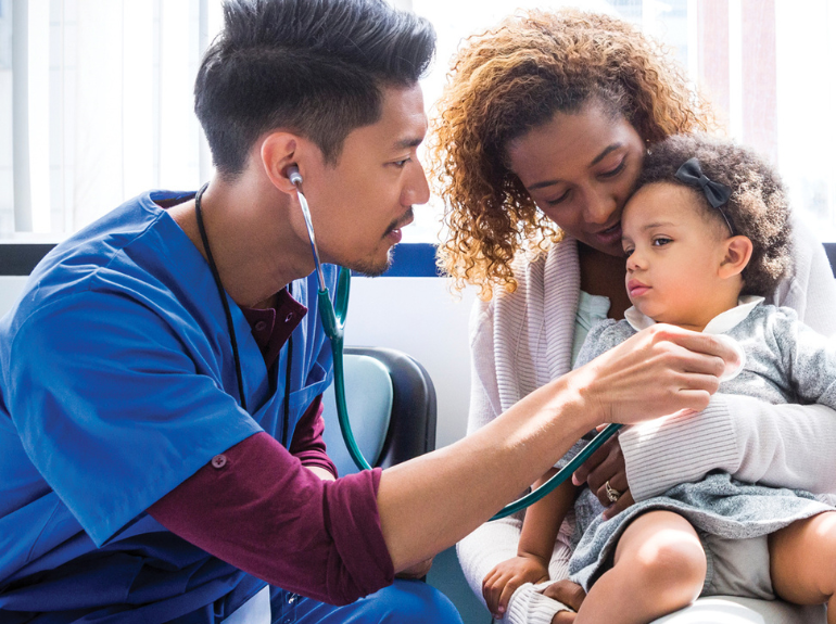 A male nurses checks the heartbeat of a young girl in her mother's lap.